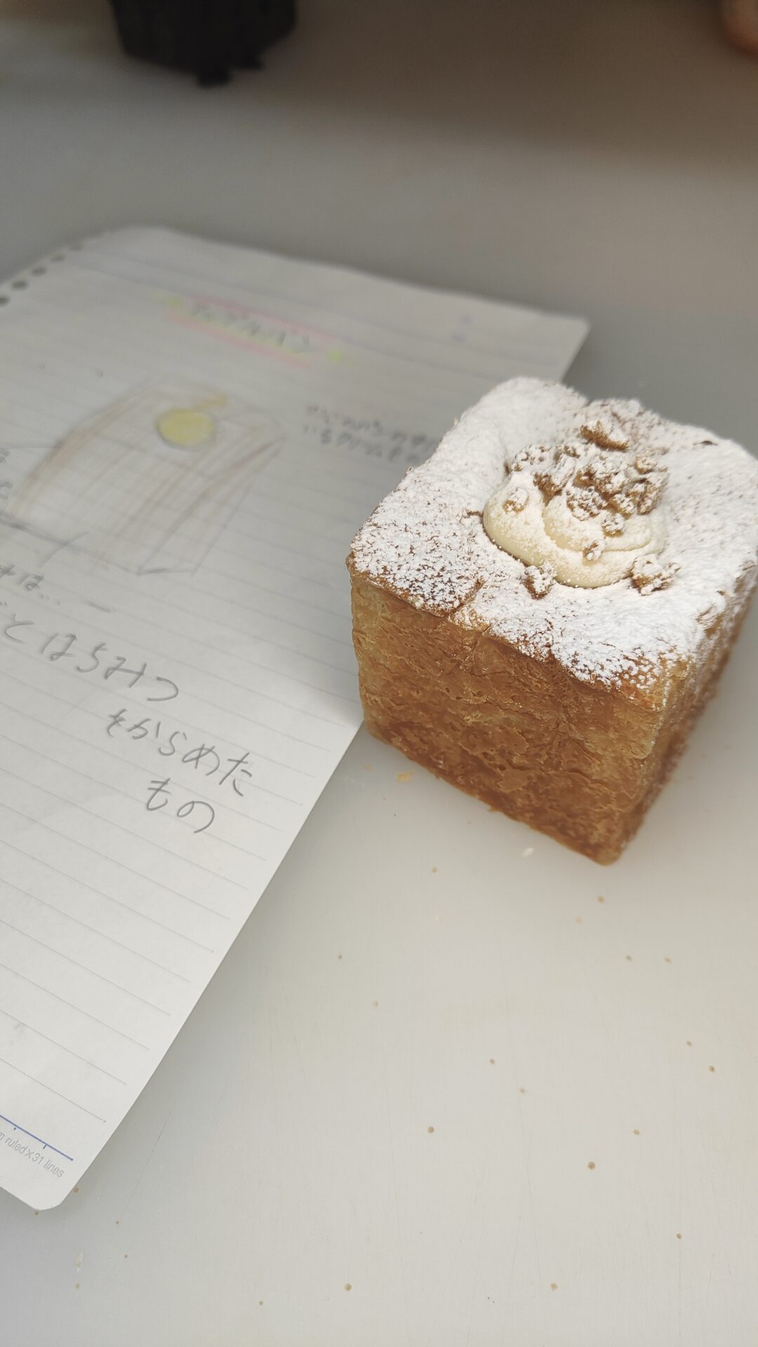 ノートに描かれた手書きのイラストと、その横に置かれた実物のキューブ型デニッシュパン。パンの表面には粉糖がまぶされ、中央にはクリームとナッツのようなトッピングが施されている。ノートには「はちみつをからめたもの」という日本語のメモがあり、商品開発やアイデア出しの様子を表現している。